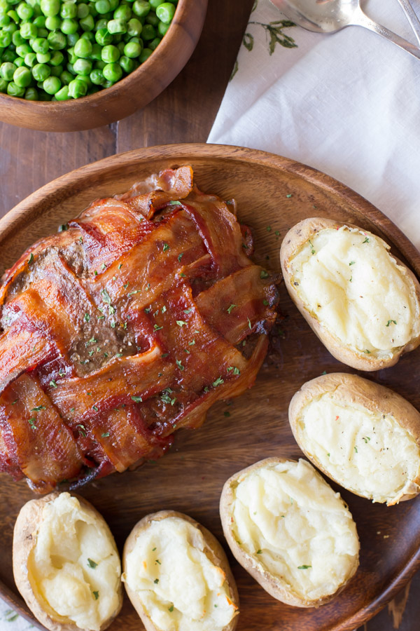 Bacon Wrapped Meatloaf on a wood serving plate with five twice baked potatoes, and a bowl of peas next to it.  