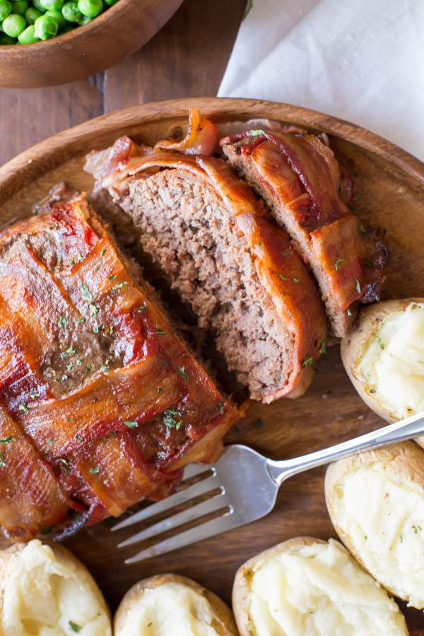 Bacon Wrapped Meatloaf that has been sliced on a wood serving plate with five twice baked potatoes and a fork, and a bowl of peas next to it.  