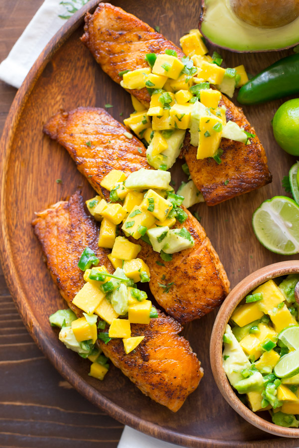 Chili Lime Salmon With Mango Avocado Salsa on a wood serving plate, with a half of an avocado, a jalapeño, a few limes, and a small wood bowl of the Mango Avocado Salsa.  