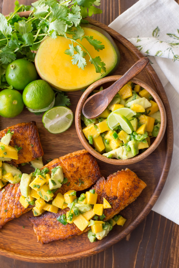 Chili Lime Salmon With Mango Avocado Salsa on a wood serving plate, with a half of a mango, a few limes, a bunch of cilantro and a small wood bowl of the Mango Avocado Salsa.  