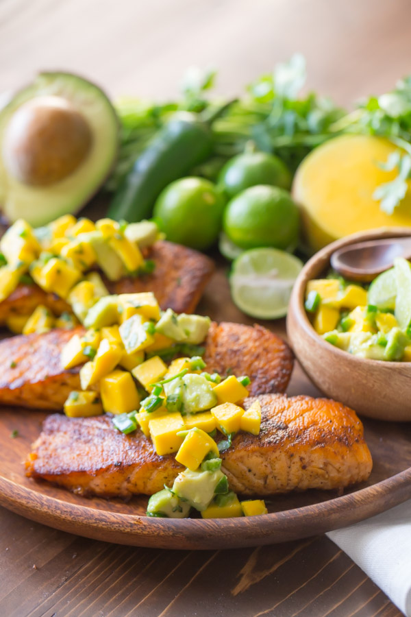 Chili Lime Salmon With Mango Avocado Salsa on a wood serving plate, with a half of an avocado, a jalapeño, a few limes, some cilantro, a half of a mango, and a small wood bowl of the Mango Avocado Salsa.  