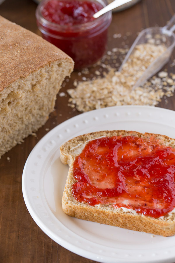 A slice of Easy Honey Whole Wheat Bread with butter and jelly on a plate, sitting next to the loaf of bread, the jar of jelly and some oats. 