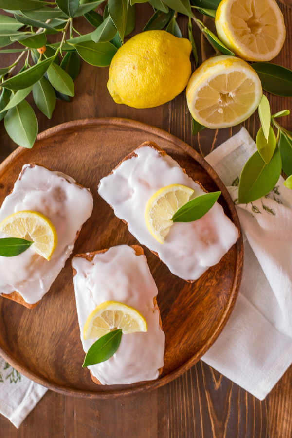 Mini Iced Lemon Pound Cake Loaves garnished with a lemon wedge and green leaf on a wood serving tray, next to some lemons and lemon tree leaves.  