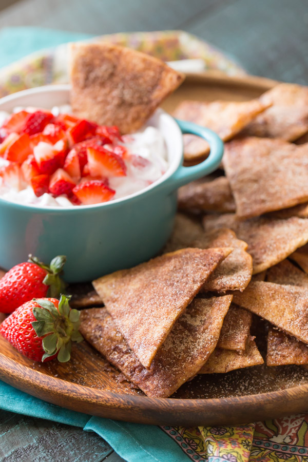 Baked Cinnamon Crisps on a wood plate with a bowl of Creamy Strawberry Dip and a few whole strawberries.  