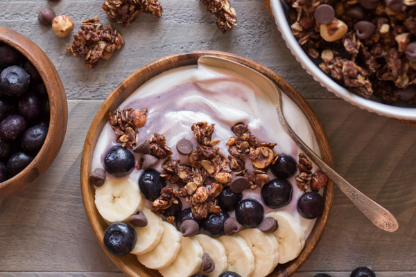 Chocolate Hazelnut Granola Yogurt Bowl with a spoon in it, and a bowl of blueberries and a bowl of granola next to the yogurt bowl. 