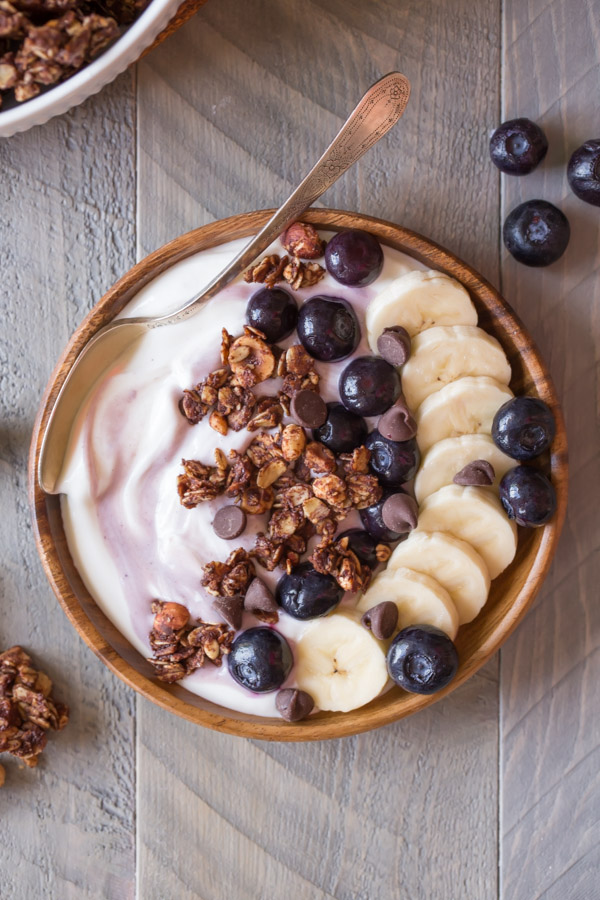 Chocolate Hazelnut Granola Yogurt Bowl with a spoon in it, and some granola and blueberries around the bowl. 