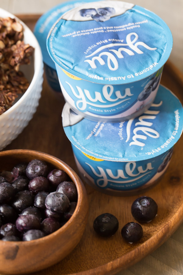 Little containers of Yulu yogurt stacked on a wood tray, along with a bowl of blueberries and and bowl of Chocolate Hazelnut Granola.