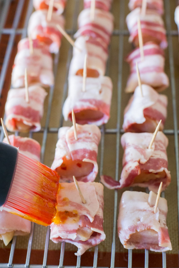 Uncooked Bacon Wrapped Chicken Bites on an oven proof rack inside a rimmed baking sheet, being brushed with Apricot Pepper Jam.  