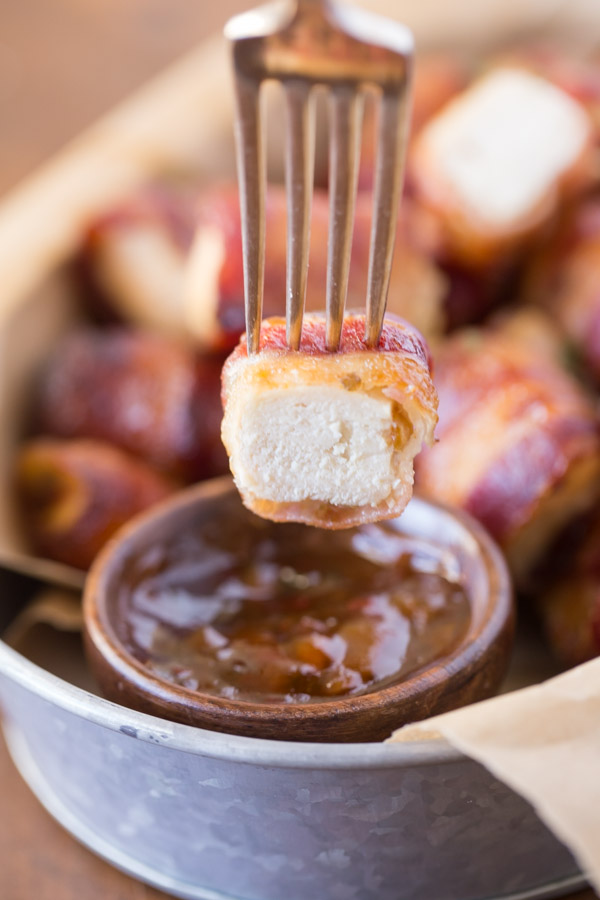A Bacon Wrapped Chicken Bite that has been cut in half, on a fork about to be dipped into a small cup of Apricot Pepper Dipping Sauce.
