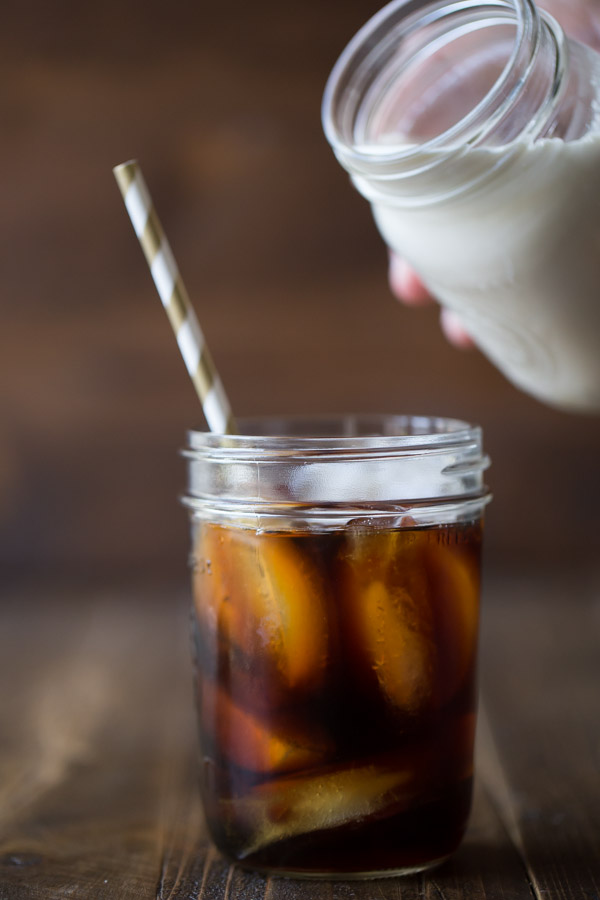 Cold Brew Iced Coffee in a glass jar with a straw, with a jar of Homemade Vanilla Creamer held over the iced coffee about to be poured in.  