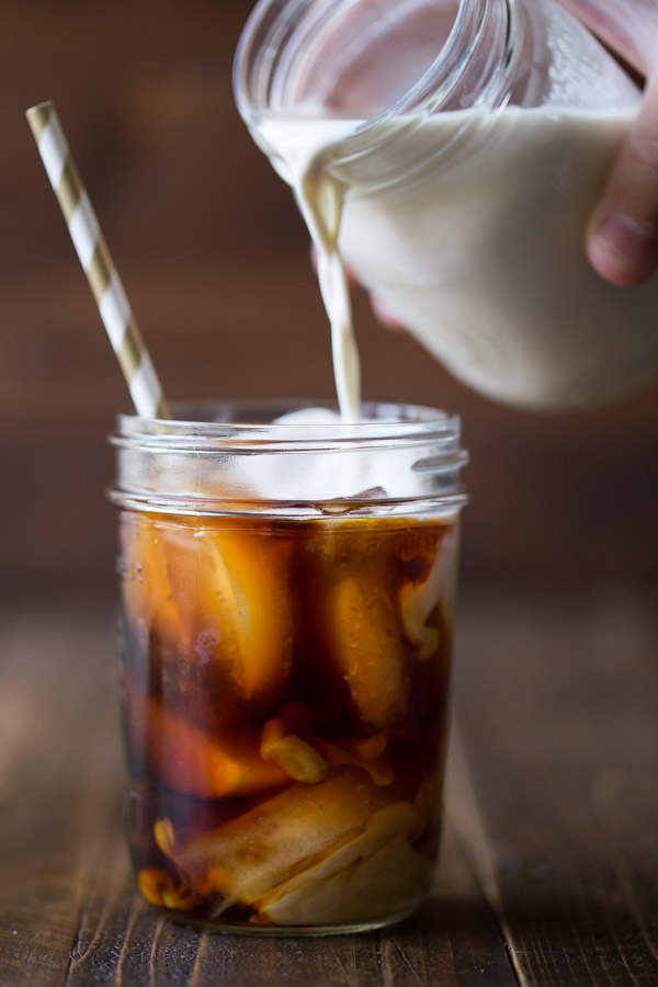 Cold Brew Iced Coffee in a glass jar with a straw, and Homemade Vanilla Creamer in a glass jar being poured into the iced coffee.  