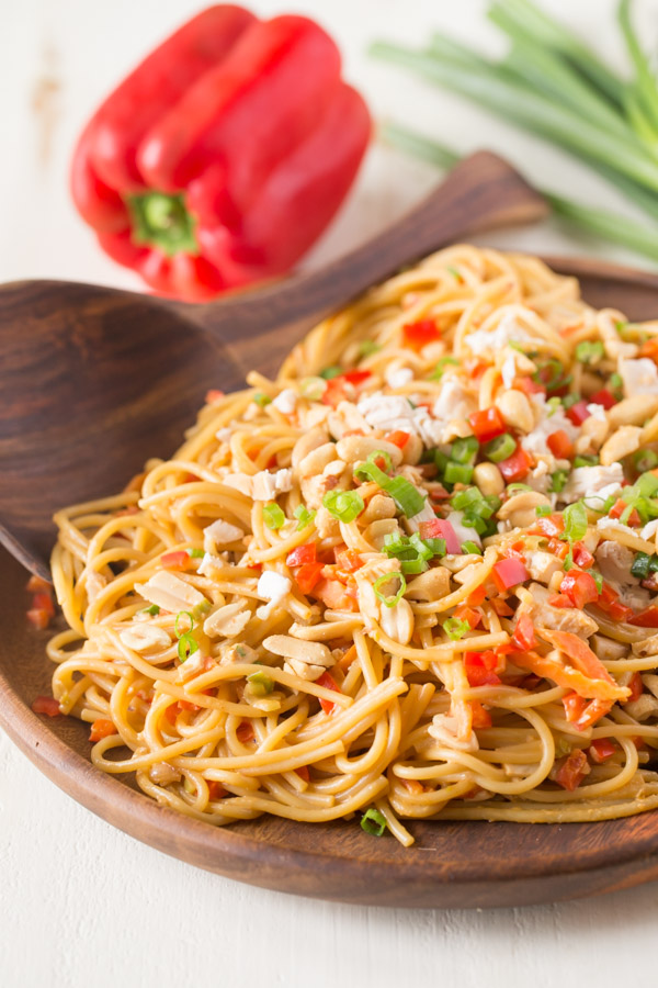 Easy Spicy Peanut Noodles With Chicken on a wood serving plate with a wood serving spoon, and a whole red pepper and a bundle of green onions in the background. 