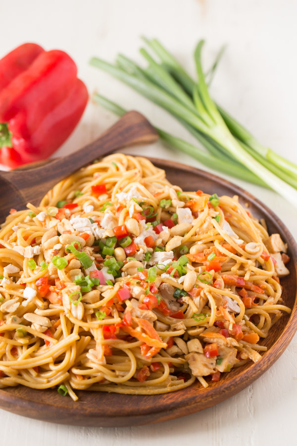 Easy Spicy Peanut Noodles With Chicken on a wood serving plate with a wood serving spoon, and a whole red pepper and a bundle of green onions in the background. 