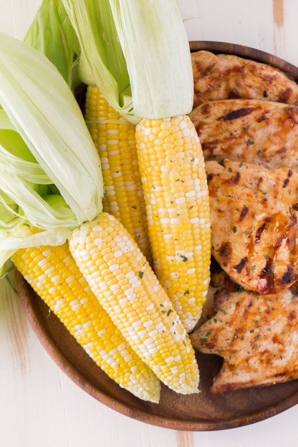 Corn on the Cob that has been cooked in the microwave, with the husks pulled back but still on the ends, sitting on a wood platter with some grilled chicken.  