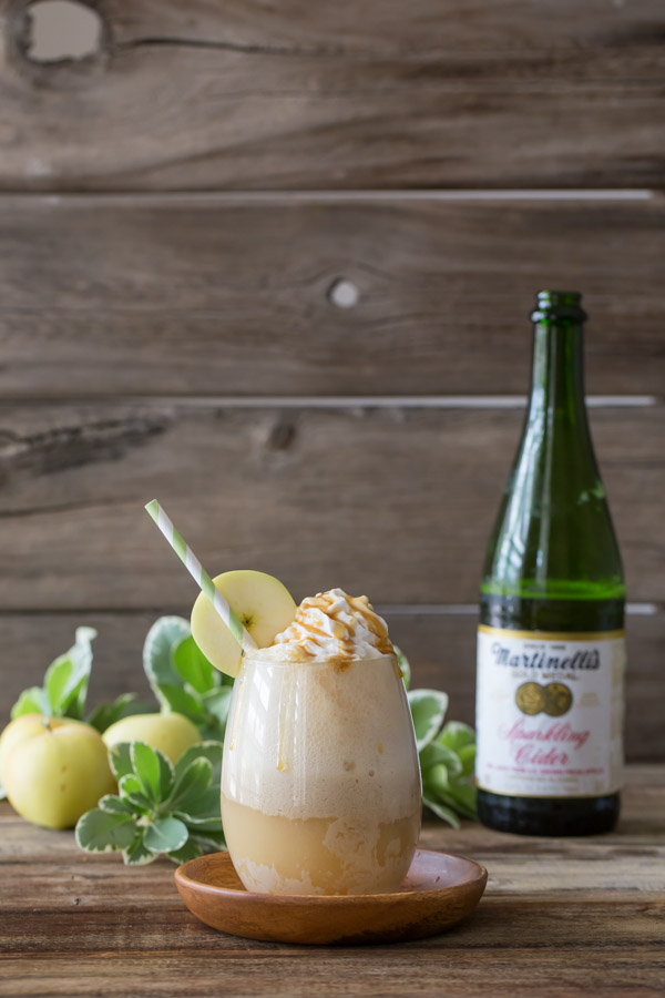 Caramel Apple Float topped with whipped cream and caramel, with a straw and an apple slice on the rim of the glass, and a bottle of Sparkling Cider and whole apples in the background.
