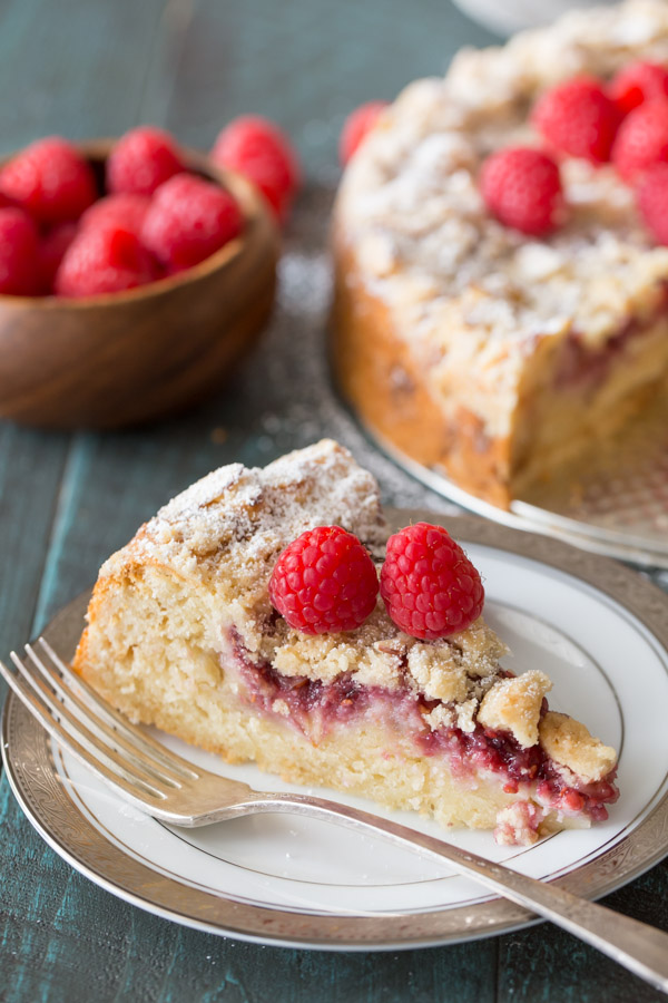 A slice of Raspberry Almond Coffee Cake garnished with powdered sugar and fresh raspberries on a plate with a fork, with the rest of the cake and a bowl of fresh raspberries in the background. 