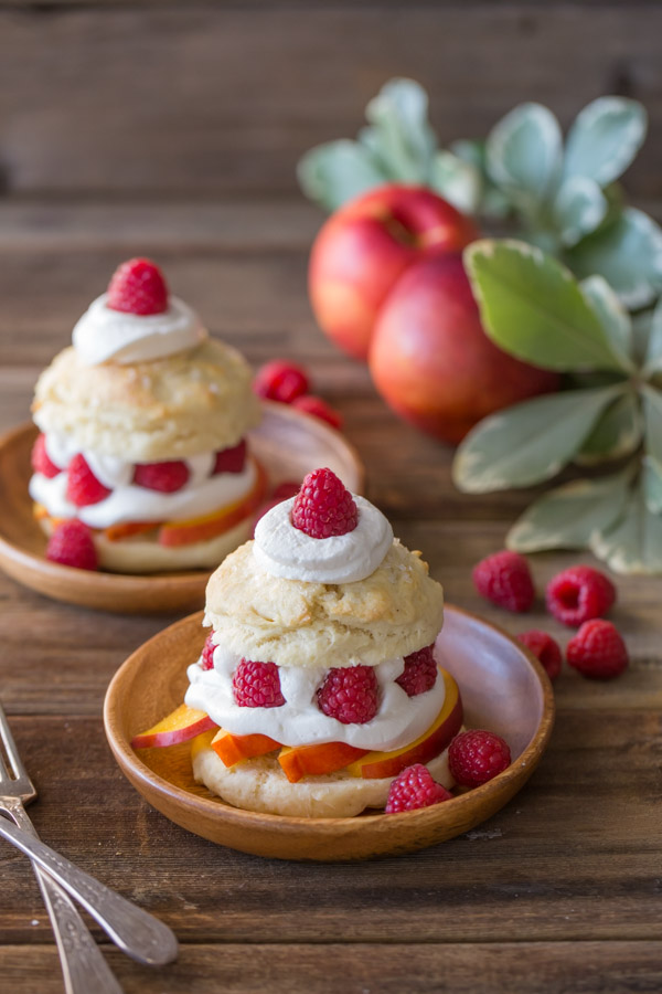 Raspberry Peach Shortcakes assembled on wood plates, with raspberries and whole peaches in the background. 
