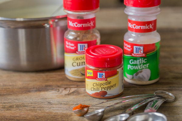 A container of McCormick Ground Cumin, a container of McCormick Chipotle Chili Pepper and a container of McCormick Garlic powder, with some measuring spoons and a sauce pan.  