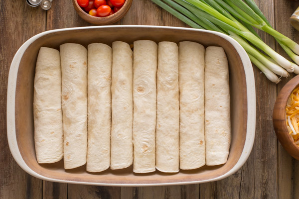 A baking dish with the stuffed, rolled tortillas in it, with other ingredients for the Breakfast Enchilada Bake next to the baking dish.  
