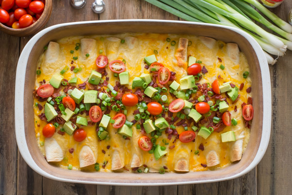 Breakfast Enchilada Bake with toppings in a baking dish, with grape tomatoes, measuring spoons, green onions, and a spice container next to the baking dish. 