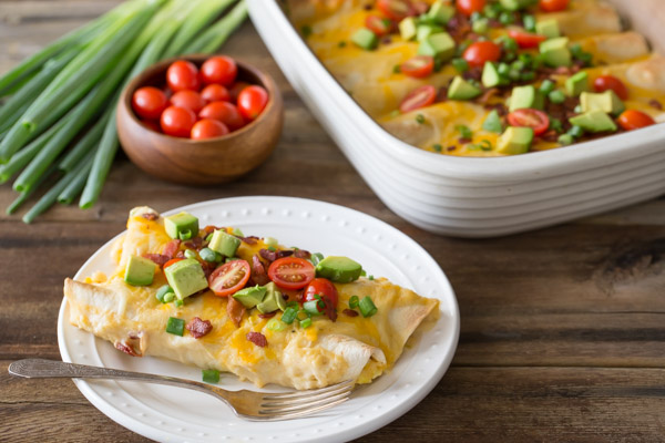 Breakfast Enchilada Bake on a plate with a fork, with the baking dish of the Breakfast Enchilada Bake in the background along with grape tomatoes and green onions.
