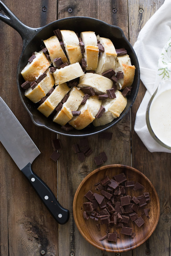 Bread and chocolate chucks arranged in a cast iron skillet, with a wood dish of chopped chocolate next to it, along with a knife and a glass cup of the cream mixture for the Chocolate Almond Bread Pudding.