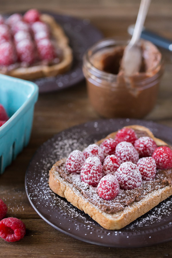 Chocolate Hazelnut Raspberry Toast sprinkled with powdered sugar on a plate, with a jar of Chocolate Hazelnut Butter in the background along with another plate of Chocolate Hazelnut Raspberry Toast sprinkled with powdered sugar.