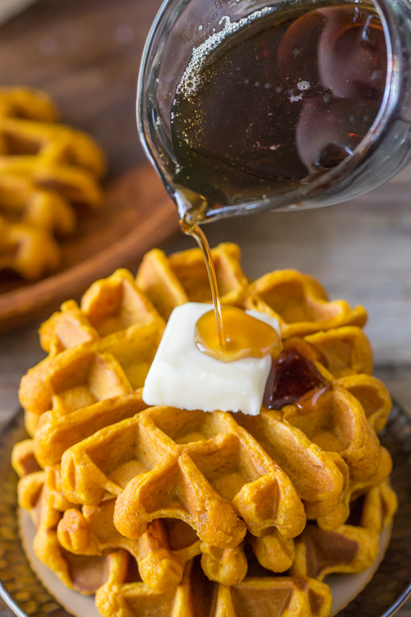 Pumpkin Spice Waffles stacked on a plate and topped with butter, with maple syrup being poured on top.  