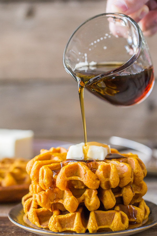 Pumpkin Spice Waffles stacked on a plate and topped with butter, with maple syrup being poured on top.  