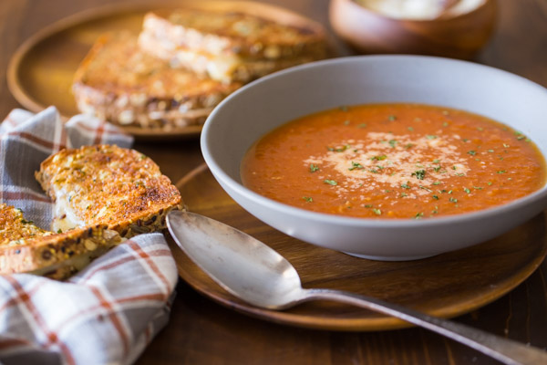 Homemade Tomato Soup in a bowl topped with grated Parmesan cheese, with a Garlic Bread Toasted Cheese Sandwich next to it and more sandwiches on a wood plate in the background.    