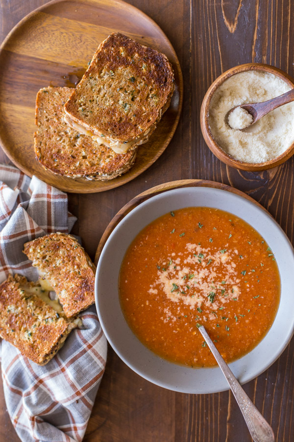 Homemade Tomato Soup in a bowl topped with grated Parmesan cheese, with a Garlic Bread Toasted Cheese Sandwich next to it, and more sandwiches on a wood plate along with a small wood bowl of grated Parmesan cheese.  