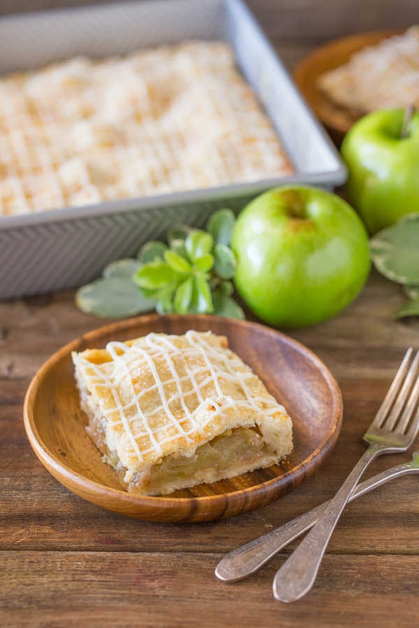 An Iced Apple Pie Bar on a wood plate, with the pan of Iced Apple Pie Bars in the background with some whole apples.  