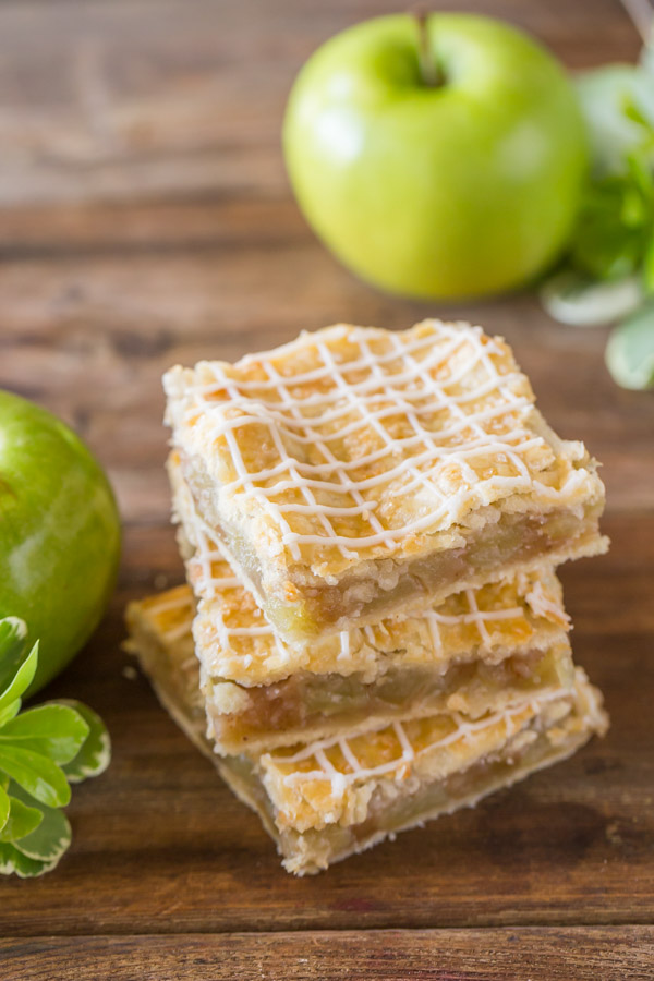 Three Iced Apple Pie Bars stacked on top of each other, with some whole apples in the background.  