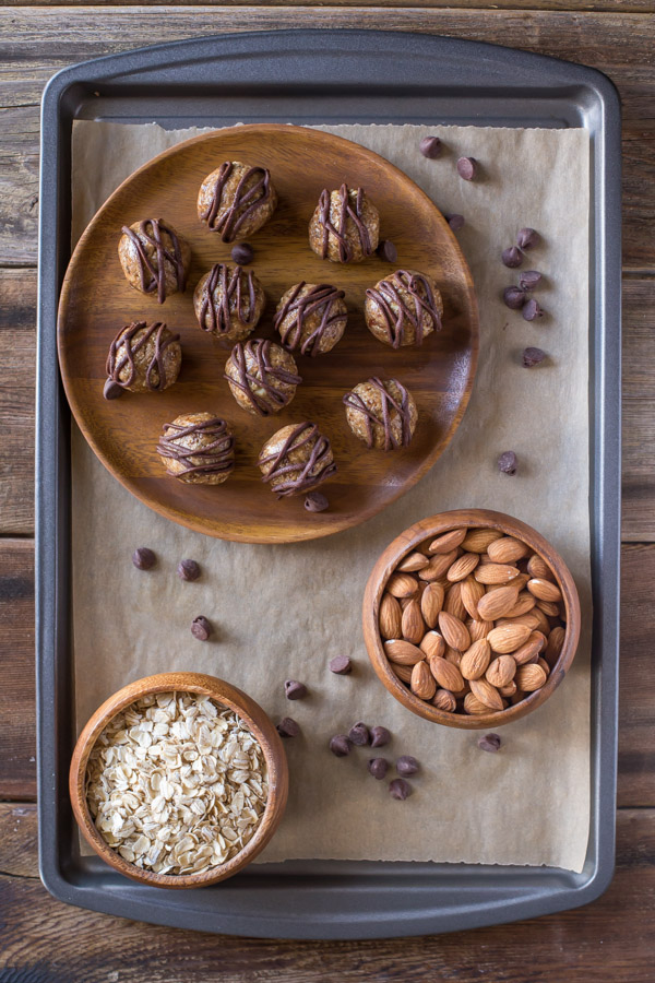 No Bake Energy Bites on a wood plate, drizzled with melted chocolate, sitting next to a bowl of almonds and a bowl of oats.  
