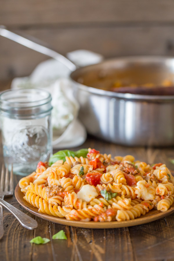 One Pot Fusilli With Tomato, Basil, and Mozzarella served on a wood plate, with a glass of water and the large pan of pasta in the background.  