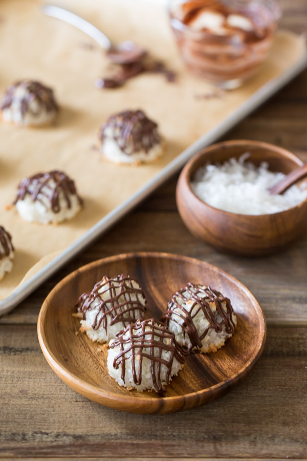 Three Stuffed Chocolate Almond Macaroons that have been drizzled with chocolate, sitting on a wood plate, with a parchment paper lined baking sheet in the background with more Stuffed Chocolate Almond Macaroons on it along with a glass cup of melted chocolate, and a small wood bowl of sweetened coconut flakes next to it.