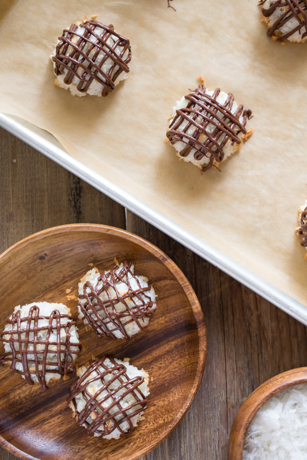 Three Stuffed Chocolate Almond Macaroons that have been drizzled with chocolate, sitting on a wood plate, next to a parchment paper lined baking sheet with more Stuffed Chocolate Almond Macaroons and a small wood bowl of sweetened coconut flakes.