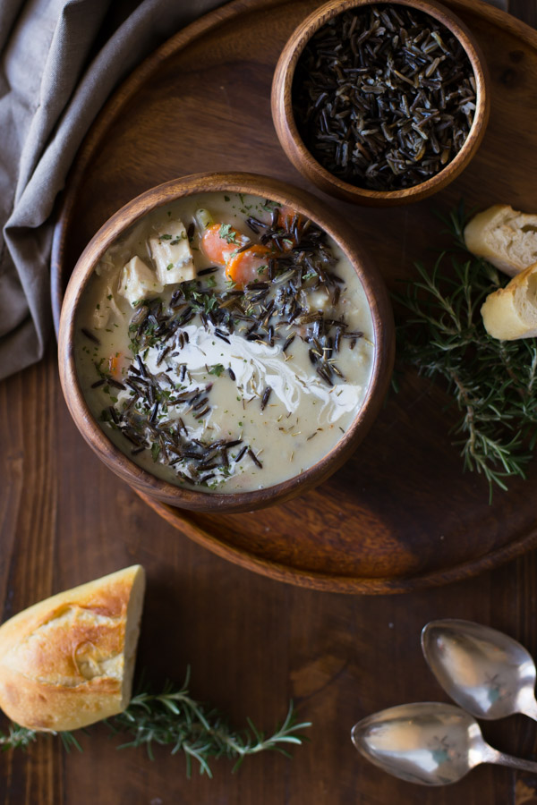 Creamy Chicken and Wild Rice Soup in a wood bowl, sitting on a wood plate along with a small wood bowl of wild rice and some bread, with two spoons and more bread sitting next to the plate. 