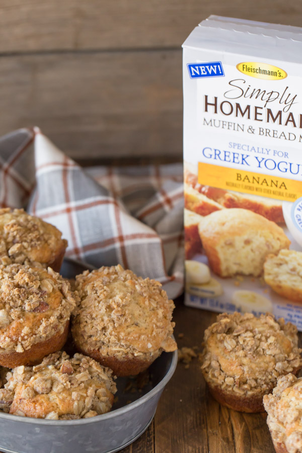 Banana Muffins With Almond Oat Streusel Topping in a galvanized bowl, sitting next to a box of Fleischmann’s Simply Homemade Muffin & Bread Mix, banana flavor and two more muffins.  