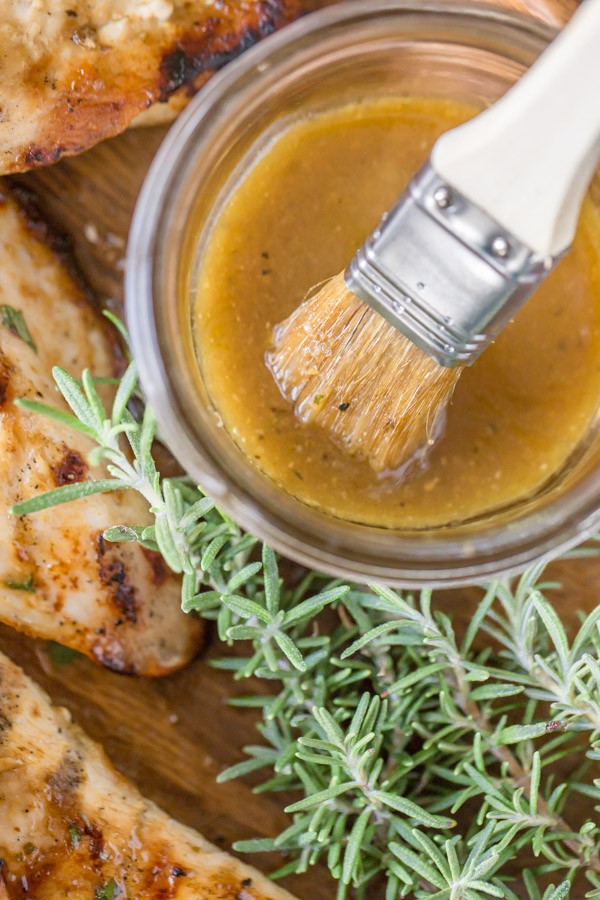 A glass jar of the Honey Mustard Sauce with a basting brush in it, sitting next to some Grilled Chicken and fresh rosemary.  