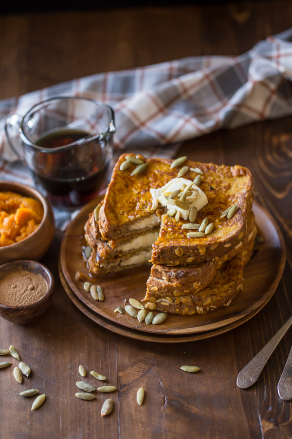 Pumpkin Pie Spiced Stuffed French Toast stacked on a wood plate and topped with butter, pepitas and maple syrup, with a section of the stack cut out.  A small wood bowl of pumpkin spice, a small wood bowl of pumpkin puree, and a glass pour cup of maple syrup are sitting next to the plate.  