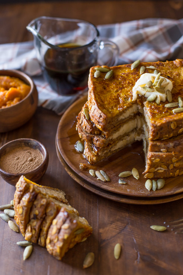 Pumpkin Pie Spiced Stuffed French Toast stacked on a wood plate and topped with butter, pepitas and maple syrup, with a section of the stack cut out and laying next to the plate, along with a small wood bowl of pumpkin spice, a small wood bowl of pumpkin puree, and a glass pour cup of maple syrup.  