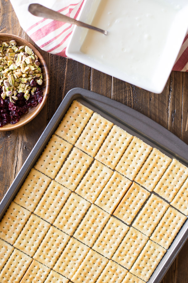 Butter crackers arranged on a baking sheet, sitting next to a wood bowl of chopped cranberries and pistachios, and a bowl of melted white chocolate.  