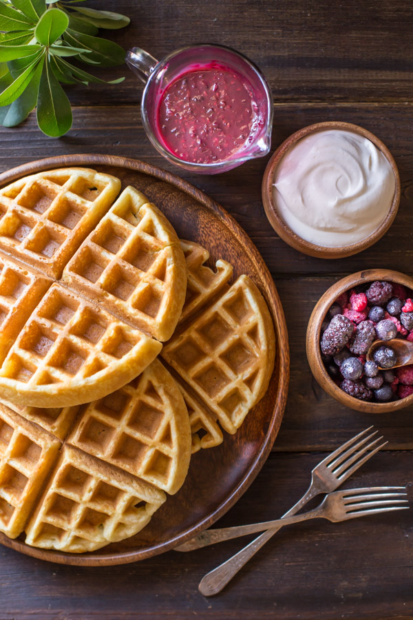 Best Ever Buttermilk Waffles stacked on a large wood plate, with a pour cup of berry sauce, a small bowl of whipped cream, a small bowl of mixed berries and two forks next to the plate. 