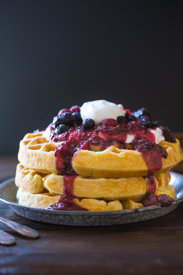 Best Ever Buttermilk Waffles stacked on a plate, topped with berry sauce, whole berries and whipped cream.  