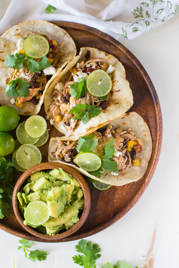 Three Crockpot Salsa Chicken Tacos arranged on a wood serving plate, with cilantro, limes and a small bowl of sliced avocado.  