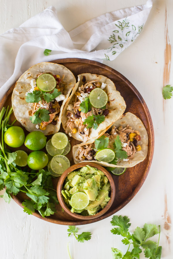 Three Crockpot Salsa Chicken Tacos arranged on a wood serving plate, with cilantro, limes and a small bowl of sliced avocado.  