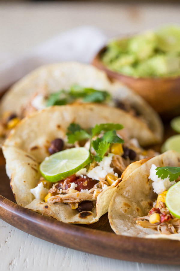 Three Crockpot Salsa Chicken Tacos arranged on a wood serving plate, with a small bowl of sliced avocado.