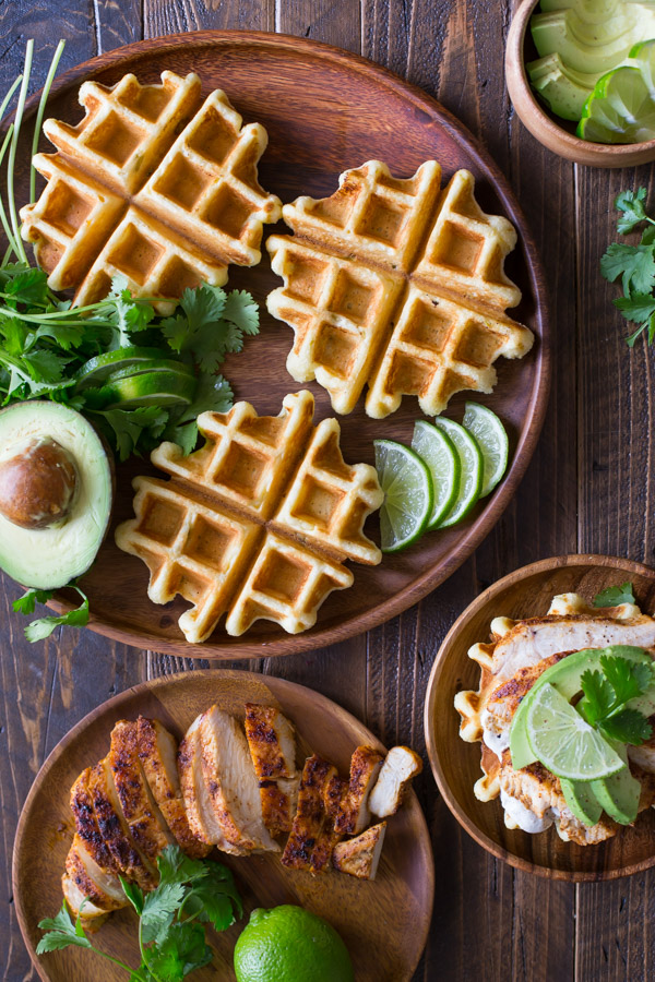 A Green Chile Chicken Waffletada assembled on a wood plate, sitting next to a large wood plate with mini cornbread waffles, lime slices, half an avocado and cilantro, a small wood bowl of avocado slices, and another wood plate with the grilled chicken on it.  