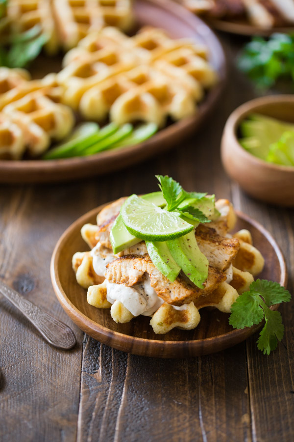 A Green Chile Chicken Waffletada assembled on a wood plate, with a large wood plate with mini cornbread waffles in the background. 